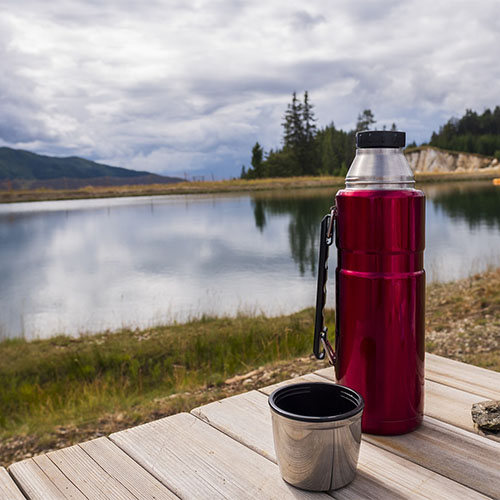 Eine rote Thermoskanne und der passende Deckel aus Edelstahl stehen auf einem Holztisch in der freien Natur. Im Hintergrund ist ein See zu sehen.