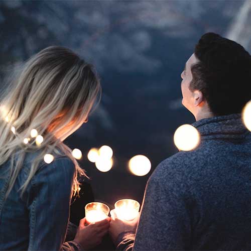 Eine Frau mit blonden Haaren und ein Mann mit dunklen Haaren sind von hinten zu sehen. Sie halten nachts Kerzen in den Händen und sind von Lichterketten umgeben, sodass eine romantische Dating-Stimmung entsteht. 
