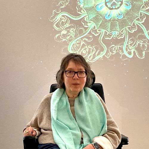 SMA-Betroffene Camilla sitzt in ihrem Elektrorollstuhl in einer Ausstellung vor einer grünen Illumination an einer weißen Wand.