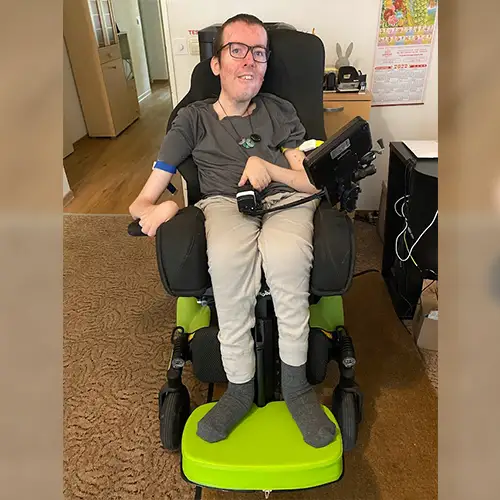 Foto des Autoren Roberto, der in seinem Rollstuhl sitzt und ein dunkelgraues T-Shirt und eine lange beige Stoffhose sowie graue Socken trägt. Das Foto wurde drinnen aufgenommen und man sieht im Hintergrund einen Wohnraum mit Regalen.