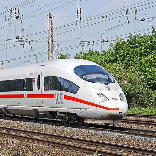 Ein Foto eines ICE-Zuges, der am helllichten Tag mit blauem Himmel unterwegs ist. Im Hintergrund befinden sich viele grüne Laubbäume, während über dem Zug zahlreiche Oberleitungsmasten entlang der Strecke zu sehen sind.