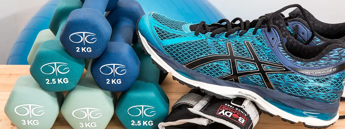 Das Bild zeigt eine Auswahl an Fitnessgeräten, darunter verschiedene Hanteln von 2 kg bis 3 kg, ein Paar türkisfarbene Sportschuhe und schwarze Gewichtsmanschetten. Im Hintergrund ist ein blauer Gymnastikball teilweise sichtbar.
