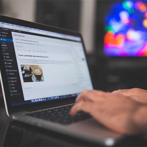 Ein Foto einer Person, die an einem Blogbeitrag auf einem Laptop arbeitet, während im Hintergrund ein bunter, verschwommener Bildschirm zu sehen ist. 