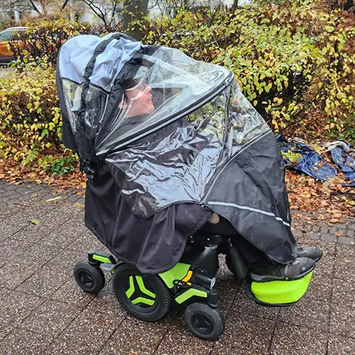 Ein Bild von Roberto, wie er in seinem elektrischen Rollstuhl sitzt, geschützt durch eine transparente Regenabdeckung, auf einem nassen Gehweg vor Büschen und Bäumen.