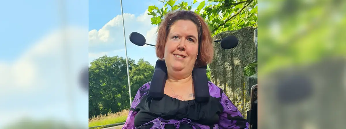 Ein Foto von Jasmin im Freien, die in ihrem Rollstuhl sitzt und freundlich in die Kamera lächelt. Sie trägt ein lila-schwarzes Oberteil und eine lange, lilafarbene Hose. Vor ihr liegt ihr selbstgeschriebenes Buch auf einem Tisch. 