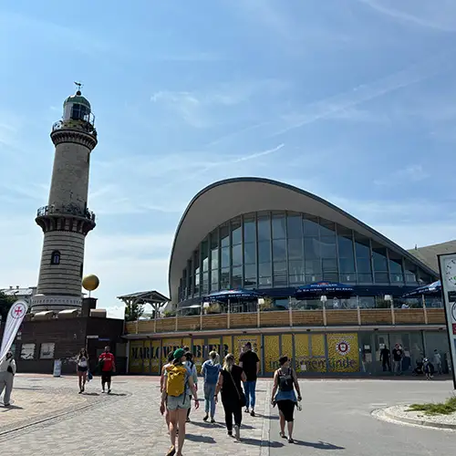 Ein Foto des Leuchtturms und der Sehenswürdigkeit „Teepott“ in Warnemünde, vor denen Menschen in Richtung der Gebäude gehen.