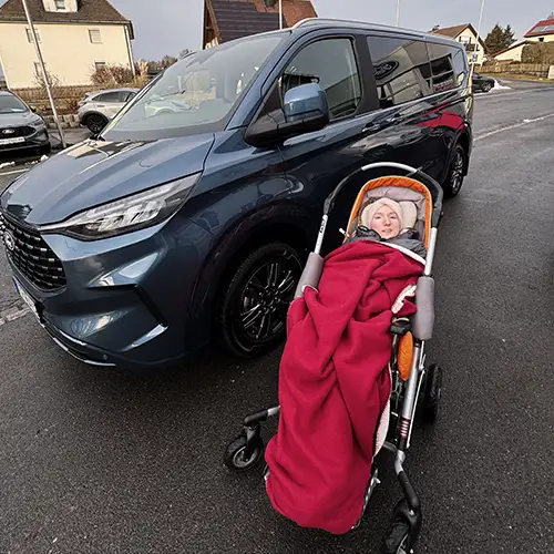 Ein Foto von Carolin, die sich draußen auf der Straße vor ihrem behindertengerechten Auto befindet und durch eine Decke gewärmt wird.
