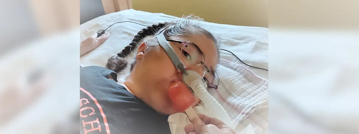 Ein Foto von Rosalie, die liegend nach vorne schaut, während sie beatmet wird und ihr ein Eis an den Mund gehalten wird.