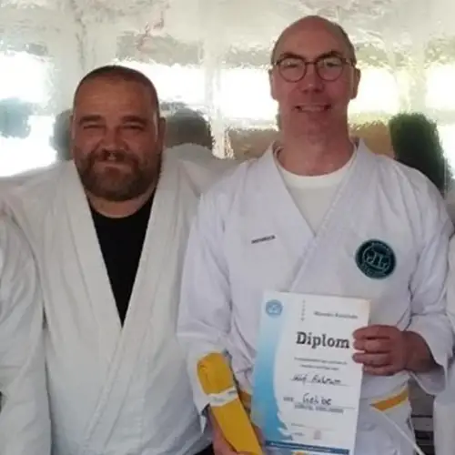Ein Foto von Ulf, der seinen gelben Gürtel und eine Urkunde in den Händen hält, während sein Trainer und er Karateanzüge tragen und nach vorne blicken.