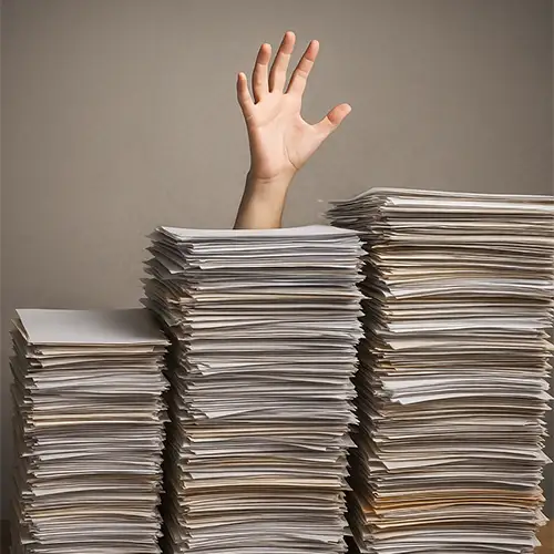 Ein Foto einer Hand, die hinter großen Papierstapeln auf einem Schreibtisch hervorragt.