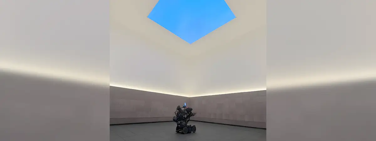 Christian sitzt in seinem eRollstuhl und betrachtet eine blaue Kunstinstallation an der Decke eines Museums. 