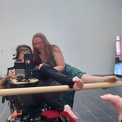 Christian sitzt in seinem eRollstuhl neben ihm lehnt seine Partnerin Grit auf ihm. Beide heben jeweils einen Fuß auf eine Ballettstange vor einem Spiegel.