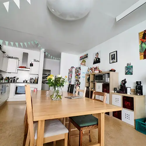Ein Bild aus Christians WG-Küche, in deren Mitte ein Holztisch mit einer Blumenvase steht. Die Wände sind dekoriert mit Bildern, Fotos und Girlanden.